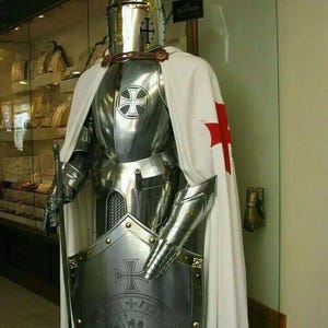 Puede incluir: Una armadura completa de plata con una capa blanca y roja. La armadura presenta un casco con visera, un peto, guantes y grebas. El caballero sostiene un escudo con una cruz y una inscripción en latín.