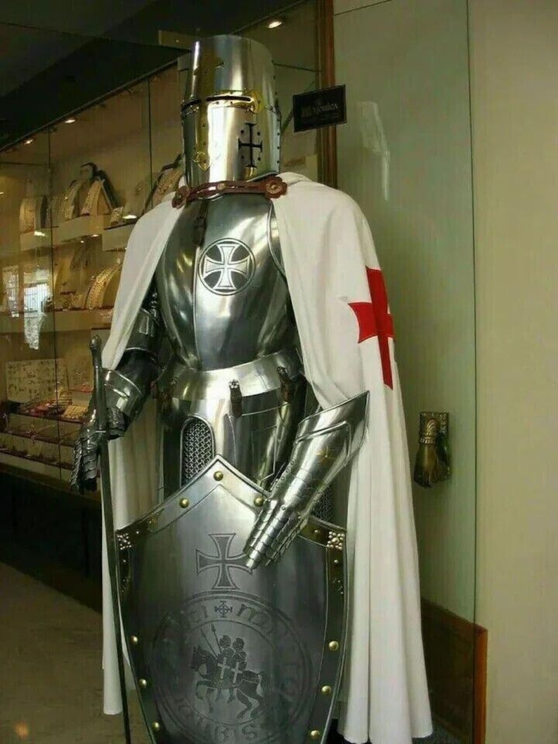 May include: A full suit of silver armor with a white and red cape. The armor features a helmet with a visor, a breastplate, gauntlets, and greaves. The knight is holding a shield with a cross and a Latin inscription.