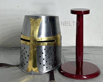May include: A silver knight helmet with a brass cross and a red wooden display stand. The helmet has a pointed top and a face mask with small holes. The stand has a round base and a tall, cylindrical post.