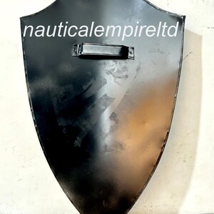 May include: A black metal shield with a handle on the top. The shield is shaped like a traditional medieval shield. The text "nauticalempireltd" is printed on the shield.