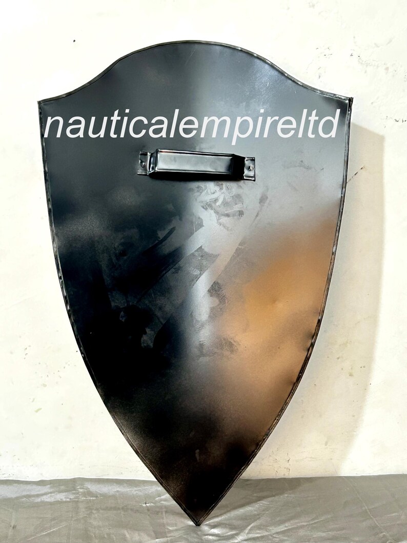 May include: A black metal shield with a handle on the top. The shield is shaped like a traditional medieval shield. The text "nauticalempireltd" is printed on the shield.