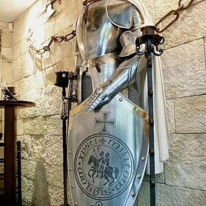 May include: A silver knight in full armor stands holding a shield with a cross and the words "In Xpsti Militom" on it. The knight is wearing a white cape and is standing on a wooden base.