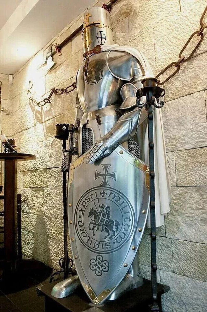 May include: A silver knight in full armor stands holding a shield with a cross and the words "In Xpsti Militom" on it. The knight is wearing a white cape and is standing on a wooden base.