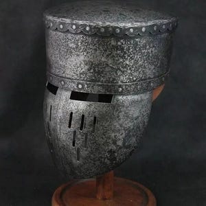 Op de afbeelding: Een zilveren ridderhelm met een vizier en een getextureerd oppervlak. De helm staat op een houten standaard.