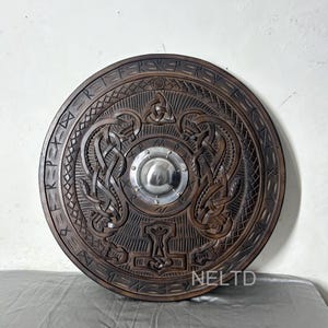 May include: A circular wooden shield with detailed carvings. The shield has a central metal boss and relief carvings of intertwined animals, runes, and a hammer symbol. The shield is stained a dark brown colour.