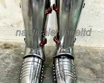 May include: A pair of silver metal shin guards with pointed toes and leather straps. The shin guards are worn over jeans.