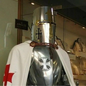 May include: A silver knight's armor with a white cape and a red cross. The knight is wearing a helmet with a gold cross on the front. The armor has a silver cross on the chest.