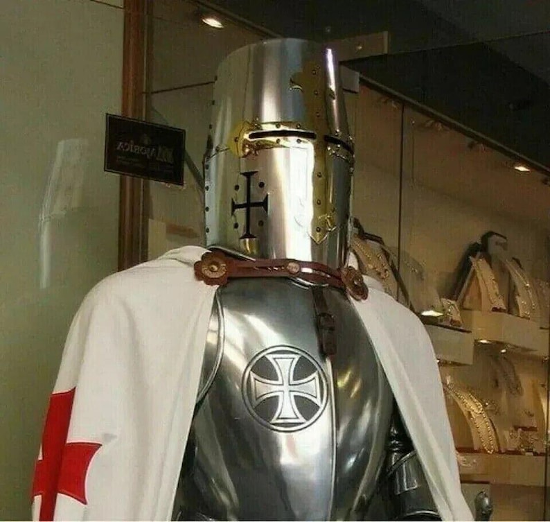 May include: A silver knight's armor with a white cape and a red cross. The knight is wearing a helmet with a gold cross on the front. The armor has a silver cross on the chest.