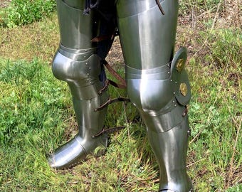 May include: A pair of silver metal leg armor with knee pads and straps. The armor is worn over brown leather pants.