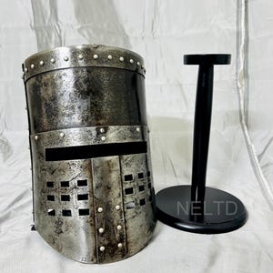 May include: A silver knight's helmet with a visor and a black wooden stand. The helmet has a textured surface and is decorated with rivets. The stand has the word "NELTD" printed on it.