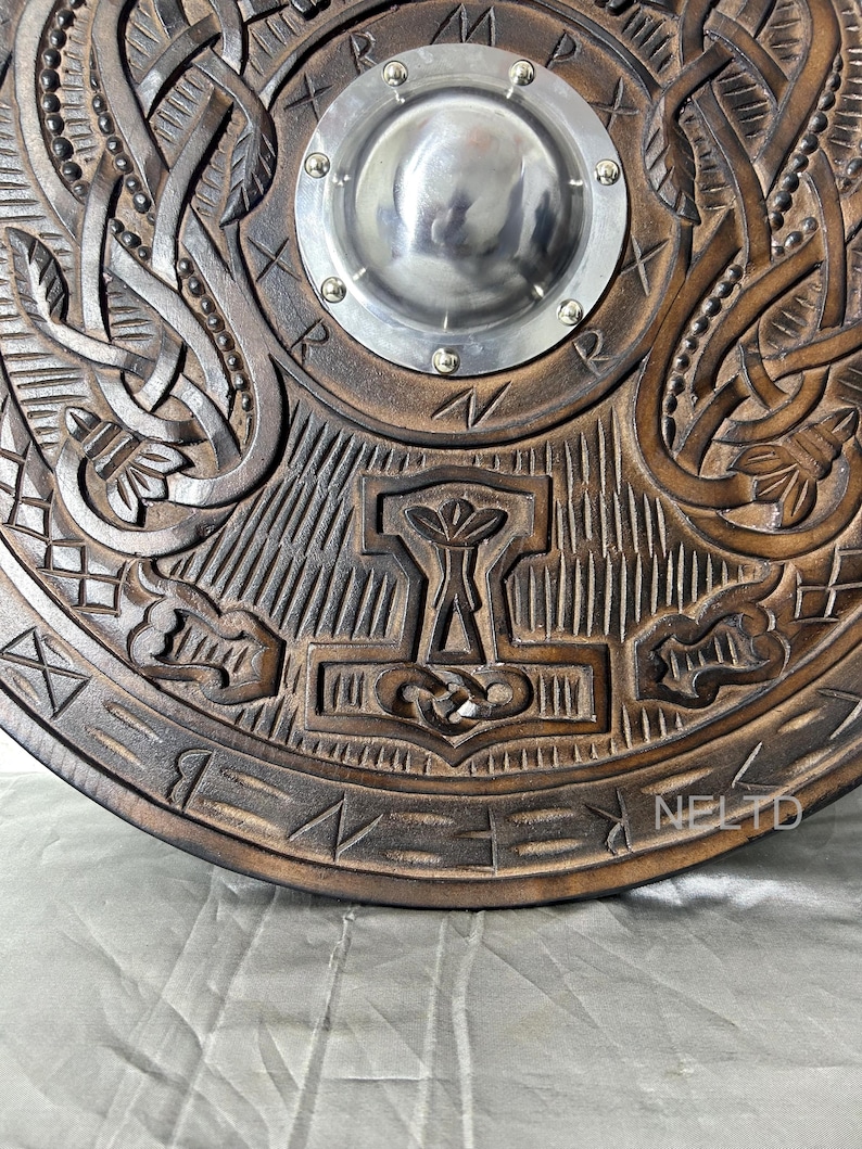 Może przedstawiać: Szczeg&oacute;łowa, ręcznie rzeźbiona drewniana tarcza z centralnym metalowym guzkiem. Tarcza posiada skomplikowane sploty i wizerunek młota Thora. Drewno ma bogate, ciemnobrązowe wykończenie, a metalowy guzik jest srebrny.