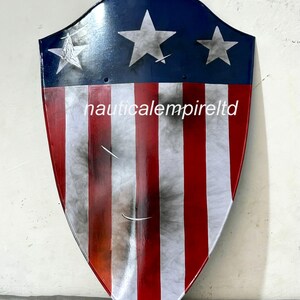 May include: A red, white, and blue shield with a distressed look. The shield has five white stars on a blue background and red and white stripes. The shield is made of metal and has a slightly curved shape. The text "nauticalempireltd" is printed on the shield.