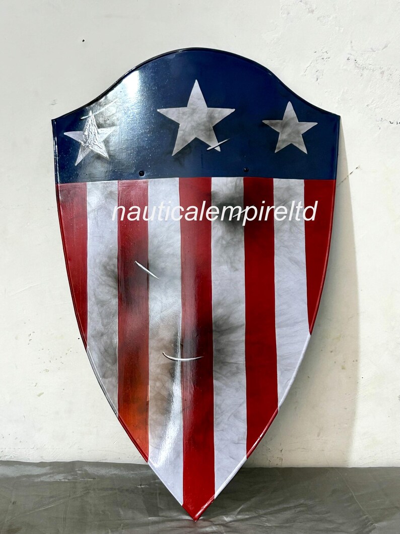 May include: A red, white, and blue shield with a distressed look. The shield has five white stars on a blue background and red and white stripes. The shield is made of metal and has a slightly curved shape. The text "nauticalempireltd" is printed on the shield.