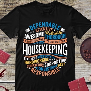 May include: A black short-sleeved t-shirt featuring a circular graphic design with the word HOUSEKEEPING in large white letters, surrounded by descriptive words including DEPENDABLE, ATTENTIVE, RELIABLE, THOROUGH, PROFESSIONAL, TRUSTWORTHY, EFFICIENT, RESPECTFUL, HARDWORKING, COURTEOUS, SUPPORTIVE, CONSISTENT, BEST, and RESPONSIBLE.
