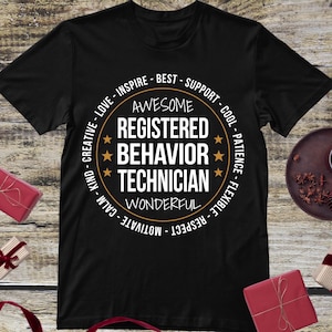 May include: Black t-shirt with a circular design featuring the words "AWESOME REGISTERED BEHAVIOR TECHNICIAN" in white. The design includes stars and words like "Inspire," "Support," and "Motivate."