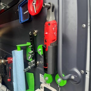 May include: A red and silver Milwaukee Fuel impact driver is mounted on a black 3D printed holder. The holder is attached to a gray surface with a green magnetic hook. A blue paper towel roll is in a green holder to the left.