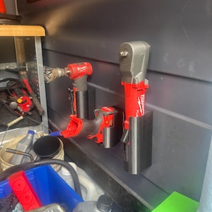 May include: Two red and black power tools are mounted on a gray wall with black plastic holders. The tools are a drill and a wrench. The tools are in a garage or workshop setting.