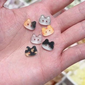 May include: Six small, cat-shaped buttons in various colors, including orange, black, gray, and white. Each button features a cat face design with two small holes for sewing. The buttons are arranged on a light-colored surface.