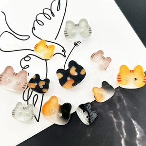 May include: A collection of cat-shaped buttons in various colors, including orange, black, gray, and brown. Each button has a buttonhole and is made of a translucent material. The buttons are arranged on a white and black surface.