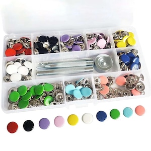 May include: A clear plastic storage container filled with assorted metal snap fasteners in red, black, purple, pink, yellow, white, blue, green, light blue, and peach colors. The kit includes two metal setting tools and a circular metal base.