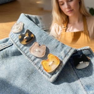 May include: A close-up of a denim jacket collar adorned with five cat-shaped buttons. The buttons vary in color, including clear, tortoiseshell, light pink, yellow-orange, and black. Each button has two holes for sewing.