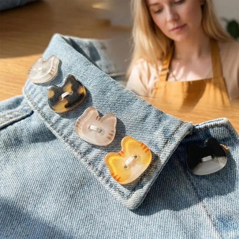 May include: A close-up of a denim jacket collar adorned with five cat-shaped buttons. The buttons vary in color, including clear, tortoiseshell, light pink, yellow-orange, and black. Each button has two holes for sewing.