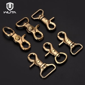May include: A collection of six gold-toned metal swivel lobster clasps arranged on a dark surface. The hardware features a spring-loaded gate mechanism and a rectangular loop for strap attachment. The WUTA logo is visible in the top left corner.