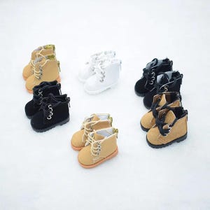 May include: A collection of miniature doll boots in various colors including tan, white, and black. Each pair features a lace-up front with metal eyelets, a side zipper closure, and a textured sole, designed for small-scale figures or display.