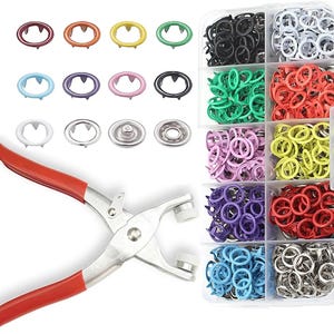 Peut inclure: Un ensemble de boutons-pression colorés dans un organisateur en plastique transparent, ainsi qu'un outil de pose de boutons-pression en métal à poignée rouge. Les boutons-pression sont de différentes couleurs, pour les projets de couture.