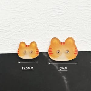 May include: Two cat-shaped buttons. The buttons are orange and white with red stripes. The smaller button is 12.5mm wide, and the larger button is 17mm wide. Each button has two holes for sewing.
