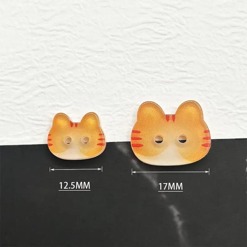 May include: Two cat-shaped buttons. The buttons are orange and white with red stripes. The smaller button is 12.5mm wide, and the larger button is 17mm wide. Each button has two holes for sewing.