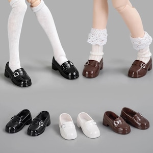 May include: A collection of miniature doll loafers in black, white, and brown, featuring a glossy finish, a rounded toe, and a small silver-toned buckle strap across the top. The shoes are displayed both on doll feet with socks and as separate pairs.