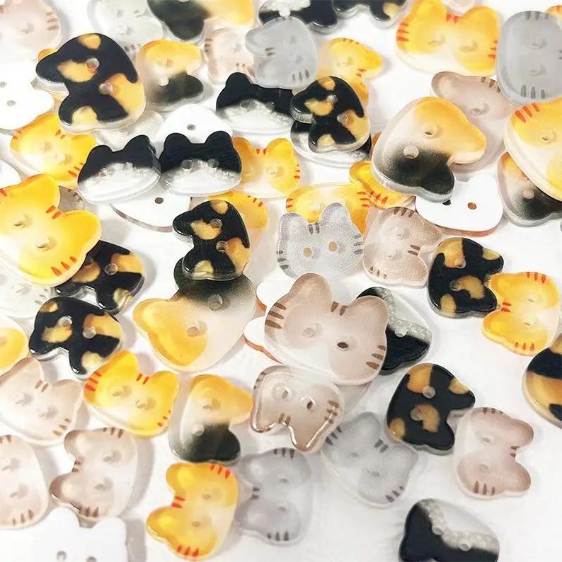 May include: A collection of cat-shaped buttons in various colors, including yellow, black, and gray. The buttons have a glossy finish and are scattered across the image. Each button features a cat face design.