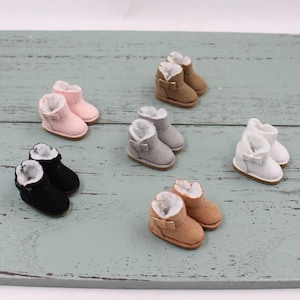 Könnte beinhalten: Eine Sammlung von Miniatur-Stiefeletten in verschiedenen Farben wie Rosa, Schwarz, Grau, Hellbraun und Weiß. Jeder kleine Stiefel verfügt über ein weißes Kunstfellfutter, eine abgerundete Zehenpartie, eine flache Sohle und eine kleine Schleife.