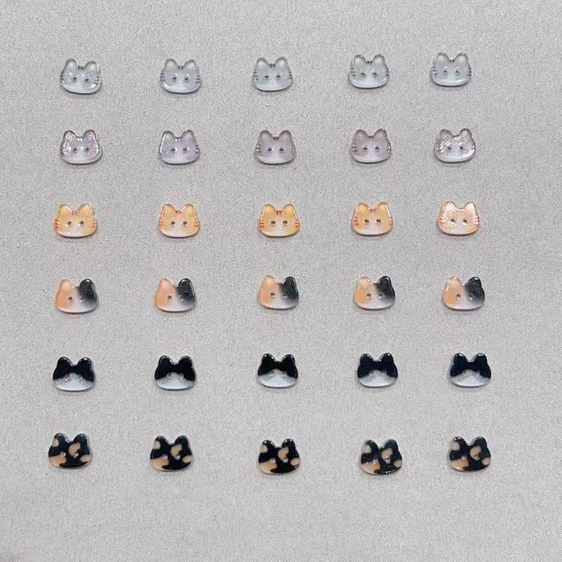 May include: A collection of twenty-five cat-shaped embellishments in various colors. The cat faces are in shades of gray, orange, black, and a tortoiseshell pattern. Each cat face has two small ears and a simple face.