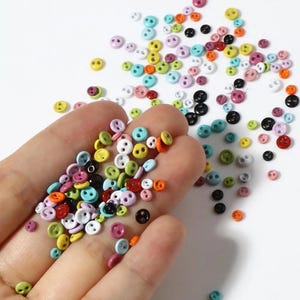 Könnte beinhalten: Eine große Sammlung winziger, runder Knöpfe mit zwei Löchern in einer Vielzahl leuchtender Farben wie Rot, Blau, Grün, Gelb, Rosa, Lila, Orange, Schwarz und Weiß, die auf einer glatten Oberfläche verstreut sind und in einer Handfläche liegen.