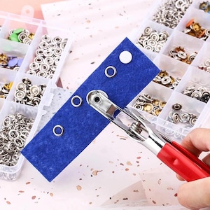 May include: A blue felt strip with metal eyelets and a white circle is held by a silver and red tool. The background shows a clear plastic organizer filled with various metal fasteners, including snaps and eyelets.