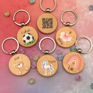 Könnte beinhalten: Sechs runde Holzschlüsselanhänger mit Metallringen. Einer zeigt einen QR-Code, die anderen Illustrationen eines Fußballs mit dem Namen Lukas, einer Ballerina mit Anna, eines Teddybären mit Emil, eines Einhorns mit Sophie und eines Regenbogens mit Lia.