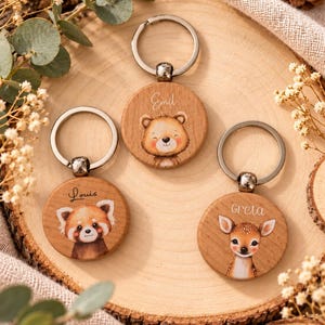 Könnte beinhalten: Drei runde Schlüsselanhänger aus Holz mit Illustrationen eines Bären, eines Roten Pandas und eines Rehkitzes. Jeder Anhänger hat einen silbernen Metallring und ist mit den Namen Emil, Louis und Greta in Schreibschrift über den Tieren versehen.