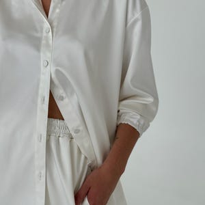 May include: A white satin shirt and matching trousers set. The shirt has a classic collar and long sleeves with button cuffs. The trousers have an elastic waistband.