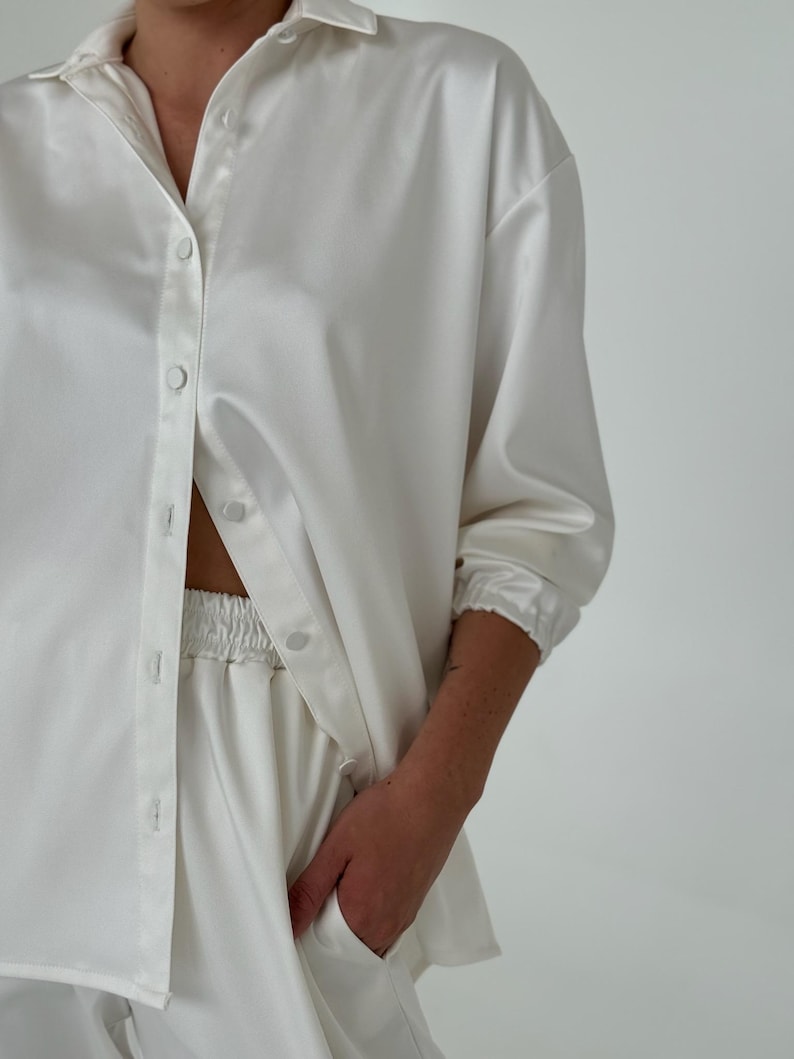 May include: A white satin shirt and matching trousers set. The shirt has a classic collar and long sleeves with button cuffs. The trousers have an elastic waistband.