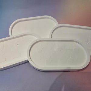Könnte beinhalten: Vier weiße, oval geformte Tabletts mit erhöhten Rändern. Zwei Tabletts zeigen Hasenmotive, eines ein Blattmuster und eines ist schlicht. Die Tabletts sind auf einem Farbverlauf aus Lila, Rosa und Blau angeordnet.