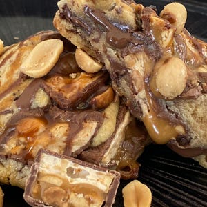 Peut inclure: Gros plan sur une barre dessert composée de couches de pâte à biscuits, de caramel, de chocolat et de cacahuètes. La barre a un aspect doux et texturé avec des noix et un filet de chocolat visibles. Le dessert est sur une assiette noire.
