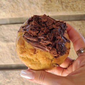 Peut inclure: Un gros cookie brun doré garni d'un tourbillon de pâte à tartiner au chocolat et d'une généreuse portion de morceaux de brownie émiettés. Le cookie est tenu dans une main, mettant en valeur sa taille et sa texture.