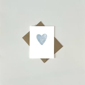 May include: A white card featuring a light blue watercolour heart design, placed on a brown envelope. The card and envelope are set against a white backdrop. The heart is centrally positioned on the card.