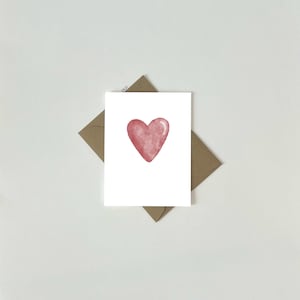May include: A white greeting card featuring a watercolour heart in shades of red. The card sits on a brown envelope, both against a white backdrop. The heart design is the focal point of the card.