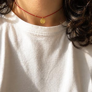 Op de afbeelding: Een verfijnde ketting bestaande uit een dun rood koord en een kleine, ronde gouden hanger met een spiraalvormig ontwerp. De hanger hangt centraal aan het koord dat rond de hals rust, wat zorgt voor een eenvoudige en strakke uitstraling.