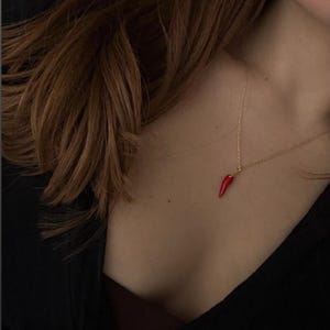 May include: A gold-coloured necklace featuring a small, red chilli pepper pendant. The pendant is a vibrant red and hangs from a delicate chain. The necklace is worn against a dark background.