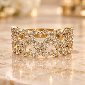 May include: A wide gold band ring featuring a repeating openwork heart pattern. The entire surface of the ring is encrusted with numerous small, clear, round-cut pavé-set stones that catch the light, creating a continuous sparkling effect.