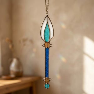 Könnte beinhalten: Ein Buntglas-Suncatcher, der an einer Metallkette hängt. Das Stück besteht aus einem spitzen oberen Teil mit weißem und türkisfarbenem Glas, einem zentralen blauen rechteckigen Paneel sowie bernsteinfarbenen und türkisen Akzenten in Metall.